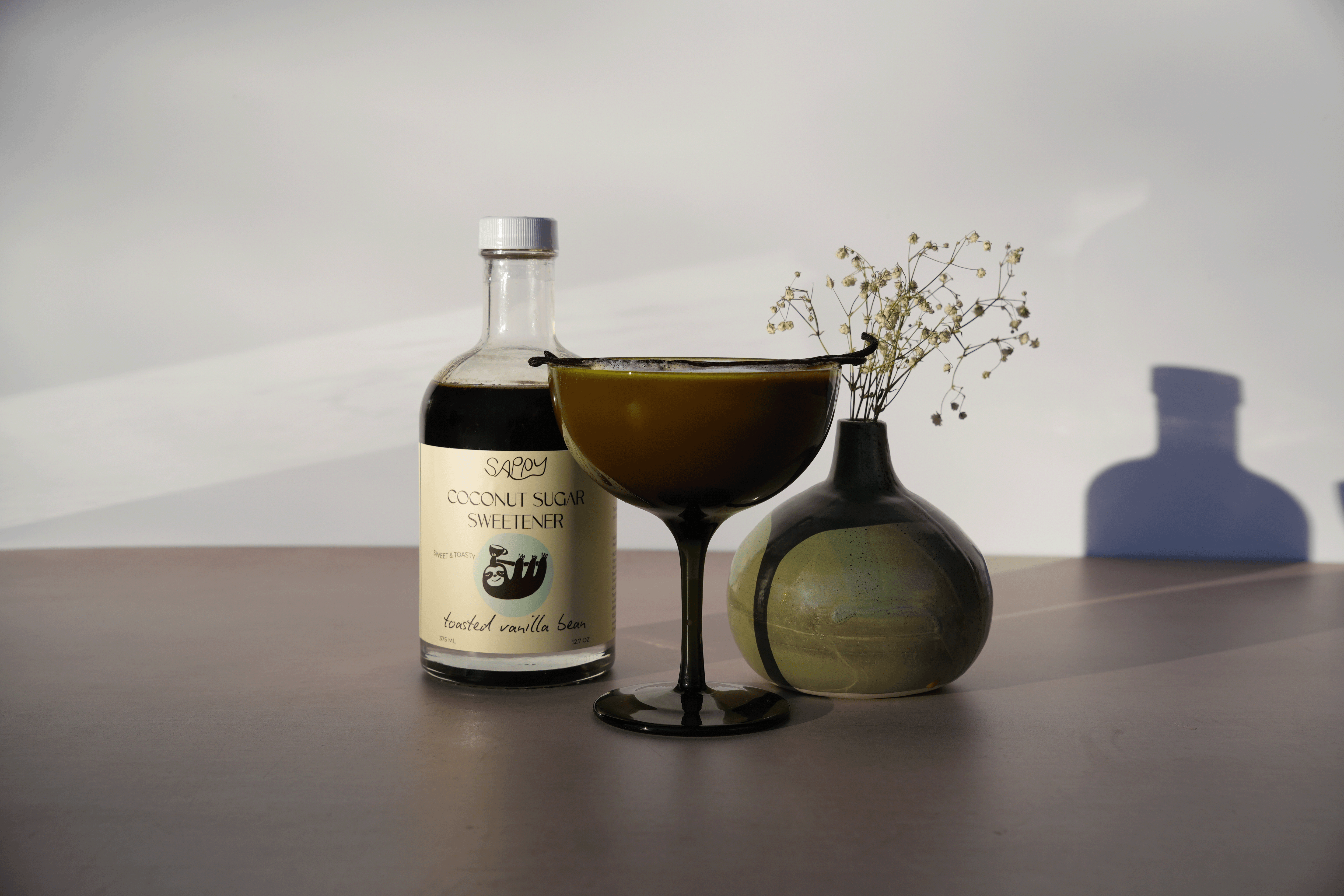 Bottle of coconut sugar sweetener, cocktail glass, and decorative vase on a table with a white background